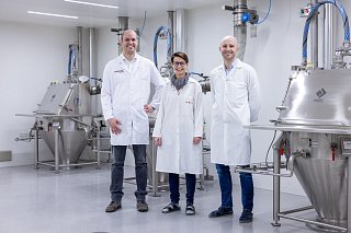 Drei Menschen in Schutzkleidung stehen lächelnd in einem Maschinenraum